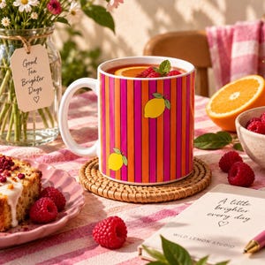 Può includere: Una tazza in ceramica bianca con un vivace motivo a strisce verticali arancioni e magenta, decorata con illustrazioni di limoni gialli. La tazza poggia su un sottobicchiere rotondo intrecciato, circondata da lamponi freschi, una fetta d'arancia e una torta.
