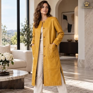 May include: A long, mustard yellow leather coat featuring a round neckline, a front button closure, and two large patch pockets. The coat has long sleeves and side slits at the hem, worn over a white top and white trousers.