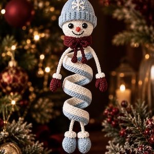May include: A crocheted snowman ornament featuring a light blue pom-pom hat with a white snowflake design, a dark red scarf, and a spiral-shaped body in alternating white and light blue stripes. The figure has dark red mittens and light blue boots.