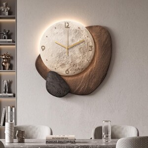 Puede incluir: Un reloj de pared con una esfera circular texturizada similar a la luna, con manecillas y números dorados en las 12, 3, 6 y 9. El reloj está montado sobre una base asimétrica de madera marrón oscura con una pieza decorativa negra y lisa.