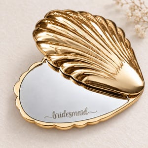 Pode incluir: Um espelho compacto dourado em forma de concha com tampa articulada. O exterior apresenta um design canelado e recortado. A superfície interna do espelho está gravada com a palavra bridesmaid em uma fonte cursiva na parte inferior.