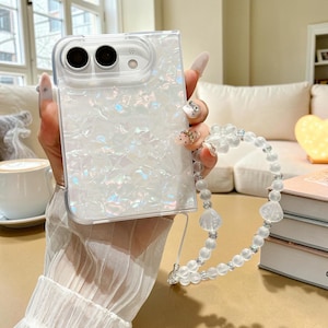 May include: A white iridescent phone case with a textured, crushed-shell appearance and a dual-lens camera cutout. Attached to the case is a beaded wrist strap featuring translucent white spherical beads and a flower-shaped charm.