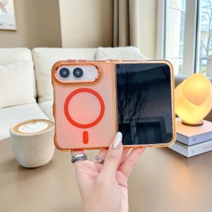 May include: A translucent orange foldable phone case featuring a prominent red circular magnetic charging ring on the back panel. The case includes a raised orange frame around the dual camera lenses and a matching orange border along the edges.