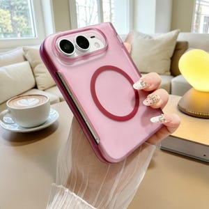May include: A translucent pink smartphone case featuring a prominent dark red circular magnetic ring on the back. The case has a raised camera cutout area with two circular lens openings and a rectangular flash cutout, designed for a mobile device.