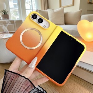 May include: A foldable smartphone case featuring a vibrant yellow-to-orange gradient finish. The back panel includes a circular magnetic charging ring and a cutout for a dual-camera system. The interior displays a black screen surface.