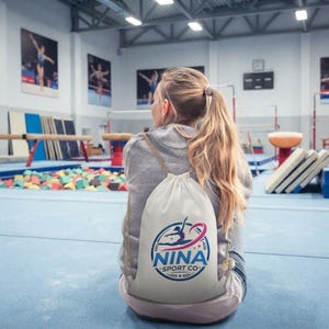 Può includere: Uno zaino a sacco bianco con un logo circolare raffigurante la silhouette di una ginnasta in blu e rosa e la scritta NINA SPORT CO. La borsa ha cordini color beige ed è indossata sopra una felpa grigia in una palestra di ginnastica al chiuso.