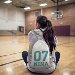 Pode incluir: Uma mochila de cordão branca com o número 07 em azul-petróleo e o nome NINA impresso numa fonte atlética arrojada. O saco tem alças finas de corda bege e é usado sobre uma camisola cinzenta clara de manga comprida.