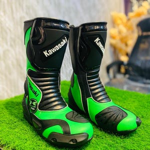 May include: A pair of Kawasaki branded motorcycle boots in black and lime green. The boots feature protective black panels on the shin and toe, accordion flex zones at the ankle, and the Kawasaki logo printed in white on the upper shaft and side.