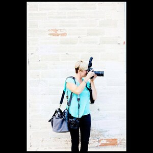 May include: A woman with short blonde hair is taking a photo with a black camera and a flash. She is wearing a turquoise shirt, black pants, and a black camera bag.