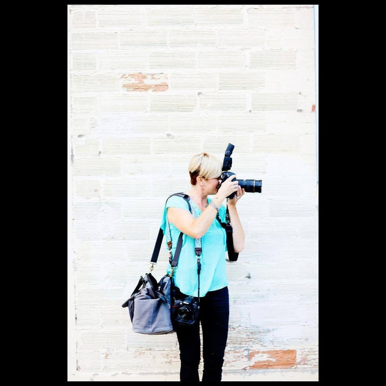 May include: A woman with short blonde hair is taking a photo with a black camera and a flash. She is wearing a turquoise shirt, black pants, and a black camera bag.