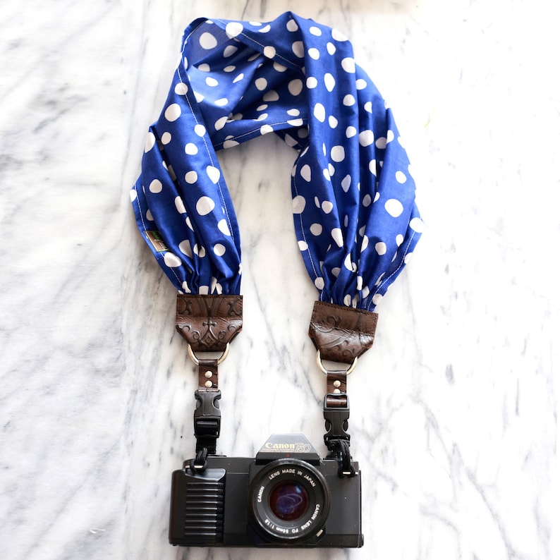 May include: A blue and white polka dot camera strap with brown leather accents and a black Canon camera attached.
