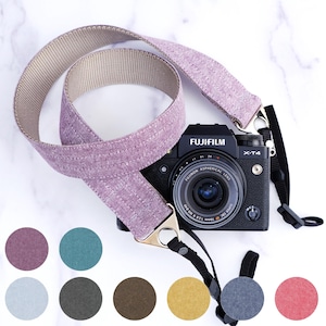 Puede incluir: Una correa de cámara de color morado y beige con un cierre de metal. La correa está hecha de un tejido tejido y tiene un aspecto texturizado. La correa se muestra con una cámara negra con el texto "FUJIFILM" y "X-T4" en ella. La cámara tiene una lente con el texto "FUJINON ASPHERICAL LENS" en ella. La correa está disponible en una variedad de colores, incluyendo morado, azul verdoso, azul claro, gris oscuro, marrón, amarillo, gris y rosa.