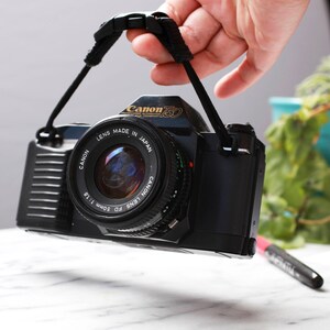 May include: A black Canon T50 film camera with a Canon FD 50mm 1:1.8 lens. The camera has a black strap attached to it.