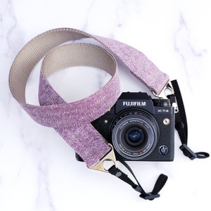 Puede incluir: Una correa de cámara de color morado y beige con una cámara negra adjunta. La cámara es una Fujifilm X-T4 con una lente Fujinon.