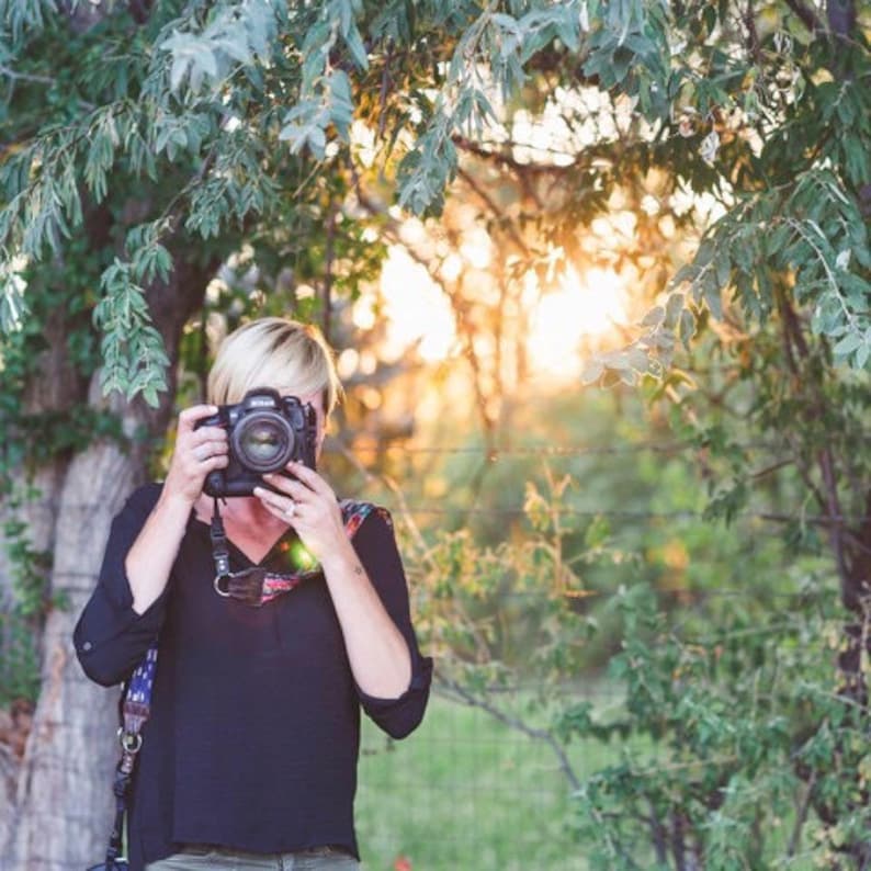 May include: A woman wearing a black shirt and a camera strap is taking a photo with a black DSLR camera. The camera is held up to her eye and she is looking through the viewfinder. The background is a blurry green and brown.