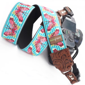May include: A turquoise and pink camera strap with a butterfly pattern and the text "MIMIGreen". The strap is attached to a brown leather camera strap with a decorative design.