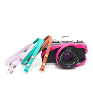 May include: A black Minolta camera with a lens, wrapped with a bright pink leather strap. Three additional leather camera straps in pastel colours: lavender, teal, and orange, are also visible. The straps have metal hardware.