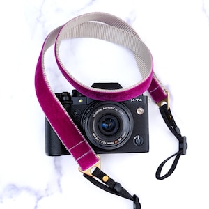 May include: A pink velvet camera strap with a gray and white striped lining. The strap is attached to a black camera with a silver lens. The camera has the text "X-T4" on the top right corner and "FUJIFILM ASPHERICAL LENS" on the lens.