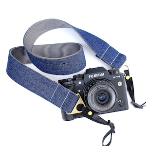 May include: A black camera with a blue and gray denim camera strap. The camera has a lens with the text "FUJIFILM" and "X-T4" on it. The lens also has the text "FUJINON ASPHERICAL LENS" on it.