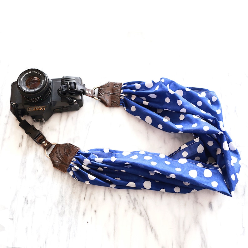 May include: A blue and white polka dot camera strap with brown leather accents. The strap is attached to a black Canon camera.