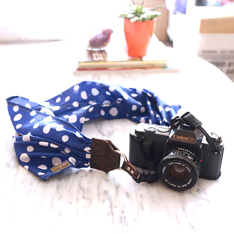 May include: A blue and white polka dot camera strap with a brown leather accent and a black Canon camera with a lens.