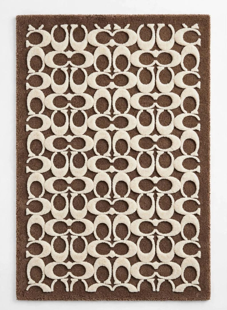 May include: A rectangular rug featuring a brown base with a repeating cream-colored pattern of interlocking C-shaped logos. The design consists of a dense, tufted texture that creates a raised, tactile surface across the entire mat.