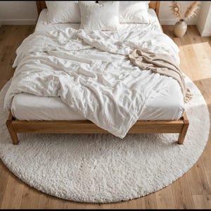 Peut inclure: Un tapis rond blanc à poils longs placé sous un cadre de lit en bois. Le lit est recouvert de linge de maison blanc, comprenant une couette, des oreillers et un jeté de lit beige plié, créant un aspect doux et superposé dans la chambre.