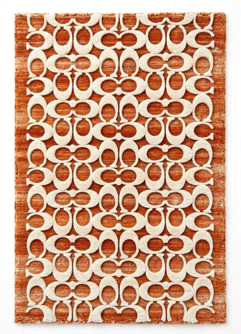 May include: A rectangular area rug featuring a repeating cream-colored C-shaped logo pattern set against a textured, burnt orange background. The rug has a thick, tufted pile construction with a consistent, symmetrical design throughout the surface.