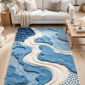 May include: A rectangular area rug featuring a textured, abstract design in shades of blue and cream. The surface displays raised, tufted patterns of varying blue tones, smooth cream-colored winding paths, and a section of small blue dots.