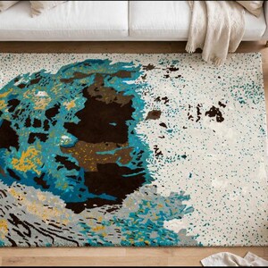 May include: A rectangular area rug featuring an abstract design with a cream-colored background speckled with teal and dark brown dots. The center displays a large, irregular shape composed of deep brown, bright teal, and golden yellow patches.