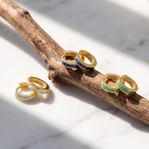 Può includere: Tre paia di piccoli orecchini a cerchio dorati con fasce di pietra intarsiate. Un paio ha pietra bianca, un paio pietra blu scuro e un paio pietra verde brillante. Gli orecchini sono disposti su un ramo di legno marrone strutturato.