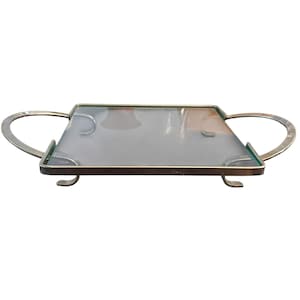 Puede incluir: Una bandeja de servicio rectangular con una superficie de cristal transparente enmarcada por un borde metálico dorado. La bandeja incluye dos asas curvas en los lados opuestos y se apoya sobre cuatro pequeñas patas metálicas a juego.