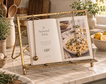 May include: A gold-toned metal cookbook stand with a distressed finish, featuring two hanging chains with weighted spheres to hold pages open. The stand displays an open book titled A Seasonal Table with text and a photograph of pasta.
