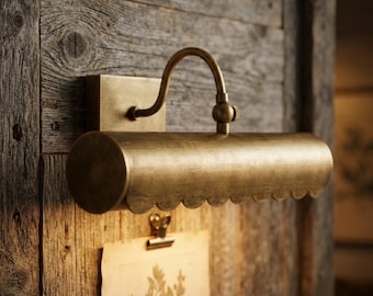 Op de afbeelding: Een wandlamp voor schilderijen met een geborstelde messing afwerking, voorzien van een cilindrische kap met een geschulpte onderrand. Het armatuur is via een gebogen arm bevestigd aan een vierkante muurplaat en schijnt naar beneden.