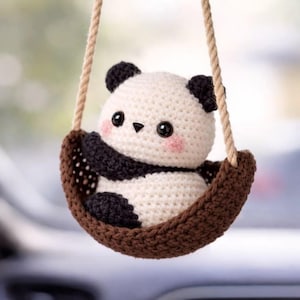 May include: A crocheted panda bear ornament with black and white yarn, featuring small black ears, black eye patches, and pink blush on its cheeks. The panda sits inside a small, brown, crocheted basket suspended by two beige twisted ropes.