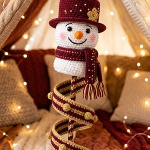 May include: A crocheted snowman ornament featuring a white head with a smiling face, orange nose, and pink cheeks. It wears a dark red hat with a gold flower and a matching scarf. A long, spiraling, striped red and gold tail hangs below.