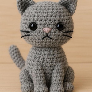 May include: A grey crocheted cat figurine featuring a round head with pointed ears, large black plastic eyes, a small pink stitched nose, and black thread whiskers. The body is seated in an upright position with a visible tail extending upwards.