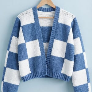 Op de afbeelding: Een gehaakt vest met lange mouwen en een blauw-wit geblokt patroon. Het kledingstuk heeft een V-hals, een knooploze opening aan de voorzijde en geribbelde boorden bij de mouwen en de zoom. Het vest hangt aan een houten kledinghanger.