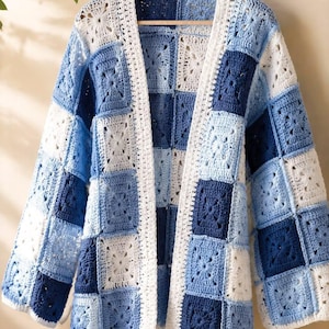 May include: A long-sleeved cardigan made from a patchwork of crocheted granny squares in varying shades of navy blue, light blue, and white. The garment features an open front design with a white crocheted border along the edges and sleeves.