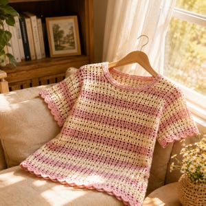 May include: A short-sleeved crochet top featuring horizontal stripes in alternating cream and soft pink colors. The garment is constructed with an open-work grid pattern and finished with a scalloped edge along the neckline, sleeves, and hem.