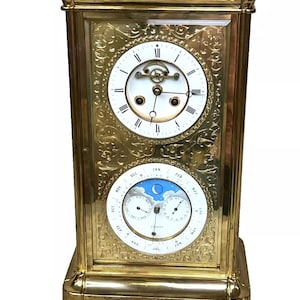 May include: A gold-toned, ornate, antique clock with a white face and Roman numeral hour markers. The clock also features a calendar dial with a blue sky and moon phase indicator.