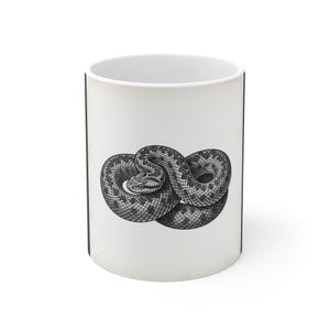 Puede incluir: Una taza de cerámica blanca con una ilustración detallada en blanco y negro de una serpiente enroscada. La serpiente está representada en un estilo realista y grabado, con escamas visibles y una mirada concentrada, centrada en el exterior.