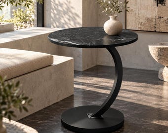 Puede incluir: Una mesa auxiliar redonda con un tablero de mármol negro con vetas blancas, sostenida por una base de metal negro curvada que se conecta a una placa base circular plana. Un pequeño jarrón beige texturizado con una planta descansa encima.