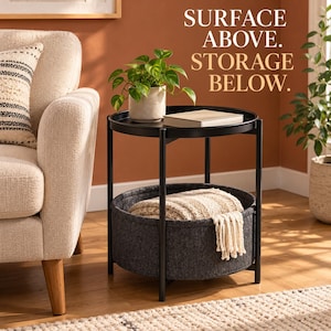 May include: A black round metal side table with a raised edge, featuring a top surface holding a potted green plant and a book, and a lower shelf containing a dark gray felt storage basket filled with a cream-colored textured throw blanket.