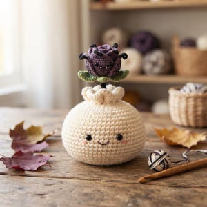 May include: A round, cream-colored crocheted plush with a smiling face and pink cheeks, topped with a dark purple crocheted rose featuring small black fangs and green leaves. The plush has a ruffled opening at the top where the flower is attached.
