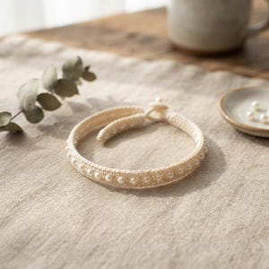 May include: A beige crocheted bracelet featuring a row of small white pearls embedded along the center. The bracelet has a circular shape with a simple loop and knot closure mechanism at the top, crafted from a textured thread material.