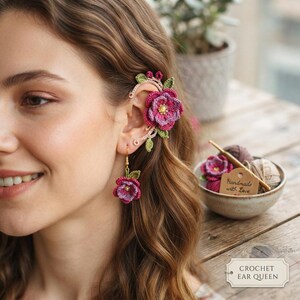 Peut inclure: Une boucle d'oreille et un ear cuff en crochet ornés de fleurs superposées violettes et roses avec des feuilles vertes. L'ear cuff entoure le haut de l'oreille, tandis que la boucle pend au lobe. Une étiquette indique "Handmade with Love."