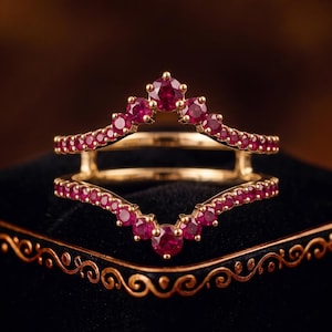 Puede incluir: Un protector de anillo de oro que presenta dos bandas en forma de cheurón conectadas por un puente. Cada banda está engastada con una serie de pequeños rubíes rojos redondos y vibrantes. El diseño crea una silueta en V, con las piedras siguiendo el contorno.
