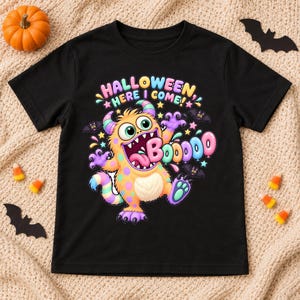 May include: A black short-sleeved t-shirt featuring a colourful cartoon monster with orange fur, purple spots, and a striped tail. The graphic includes the text "HALLOWEEN HERE I COME!" and "BOOOOO" in bubble letters, surrounded by small bats and stars.