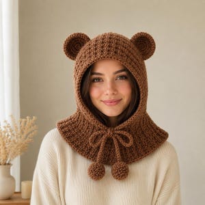May include: A brown crochet balaclava hood featuring two rounded bear ears on top. The hood is constructed with a textured stitch pattern and includes a drawstring closure at the neck with two dangling pom-poms at the ends of the ties.